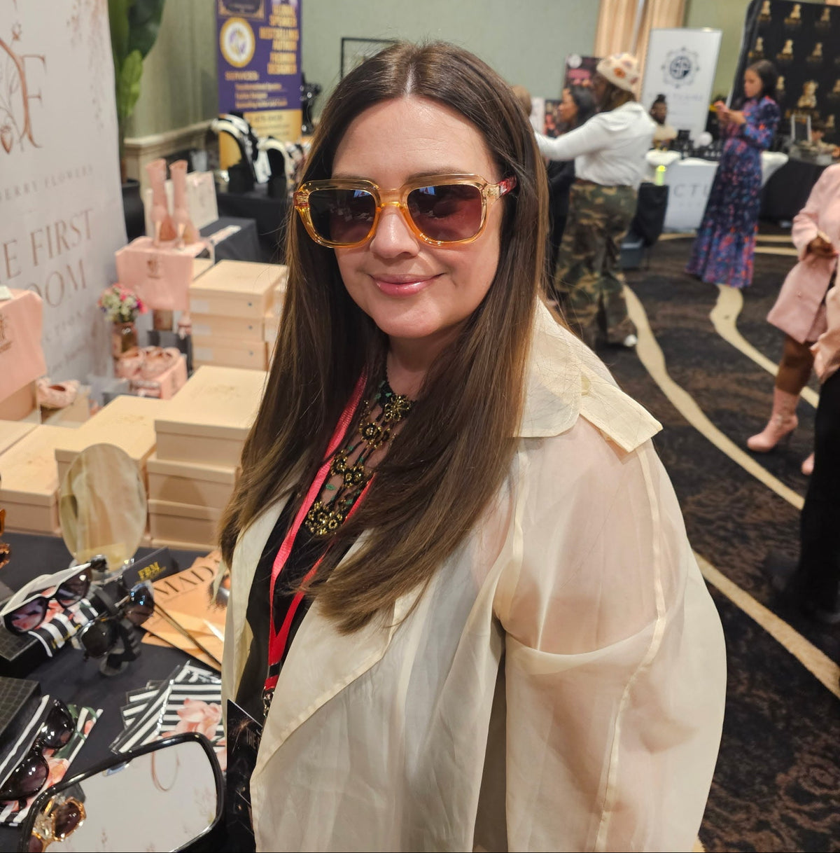 A woman with long brown hair wearing large tortoiseshell sunglasses and a cream-colored trench coat stands at a booth at an event. The booth displays pink and beige gift boxes and cosmetics, with event banners and other attendees visible in the background.