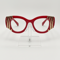 Red cat-eye eyeglasses with gold rhinestone embellishments on the temples, displayed on a clear acrylic stand against a white background.