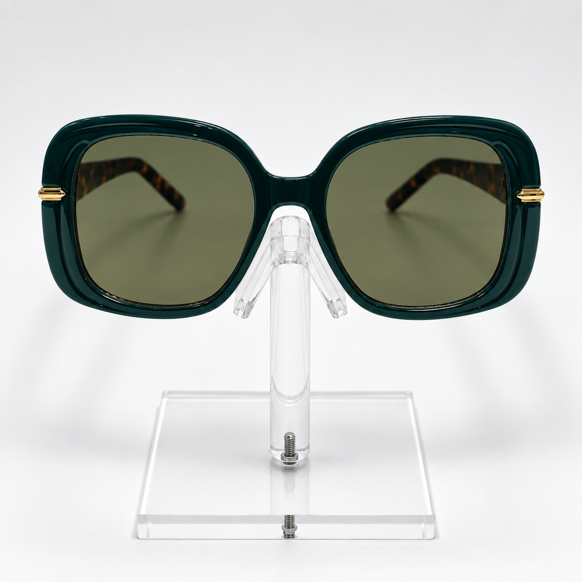 Oversized square sunglasses with a dark green frame, olive green lenses, and gold metal accents on the temples, displayed on a clear acrylic stand against a white background.