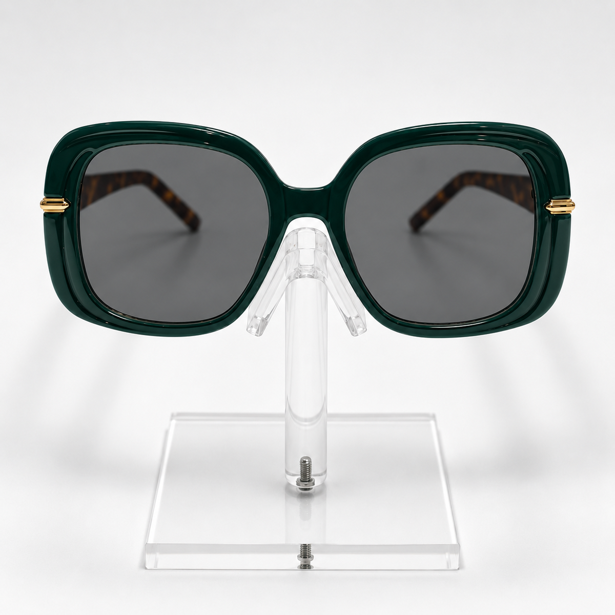 Oversized square sunglasses with a dark forest green frame, dark gray lenses, and gold metal accents on the temples, displayed on a clear acrylic stand against a white background.