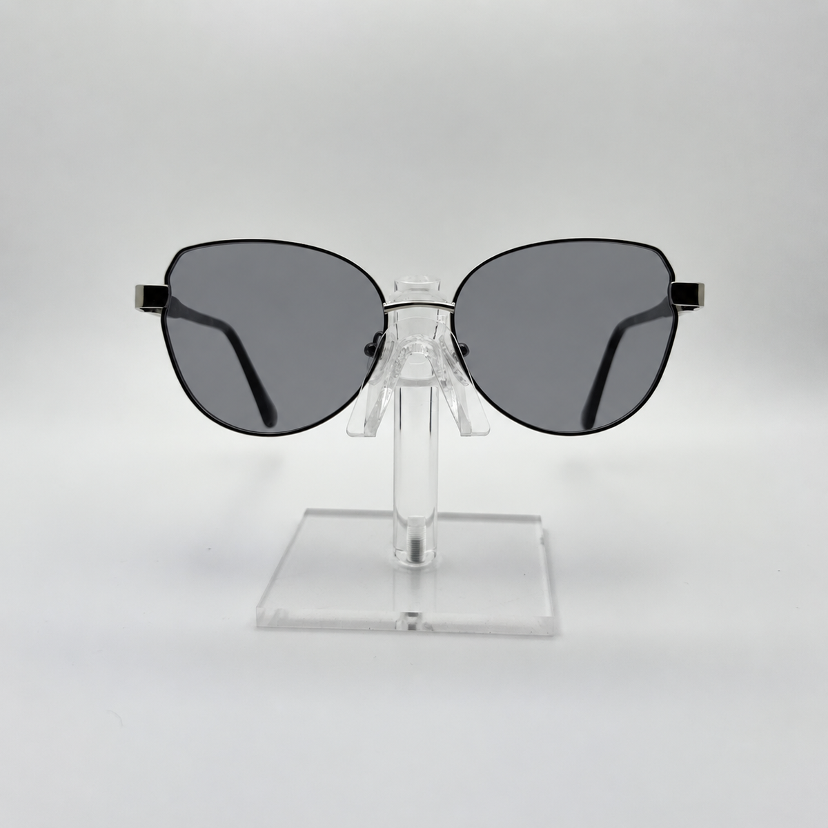 Oversized cat-eye sunglasses with dark gray lenses and black metal frames, displayed on a clear acrylic stand against a light gray background.