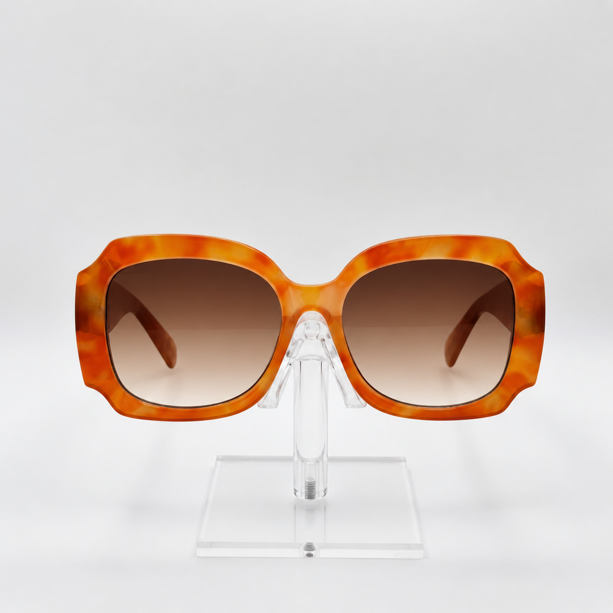 Oversized square sunglasses with a marbled orange and yellow acetate frame and brown gradient lenses, displayed on a clear acrylic stand against a light gray background.
