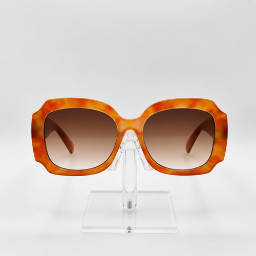 Oversized square sunglasses with a marbled orange and yellow acetate frame and brown gradient lenses, displayed on a clear acrylic stand against a light gray background.