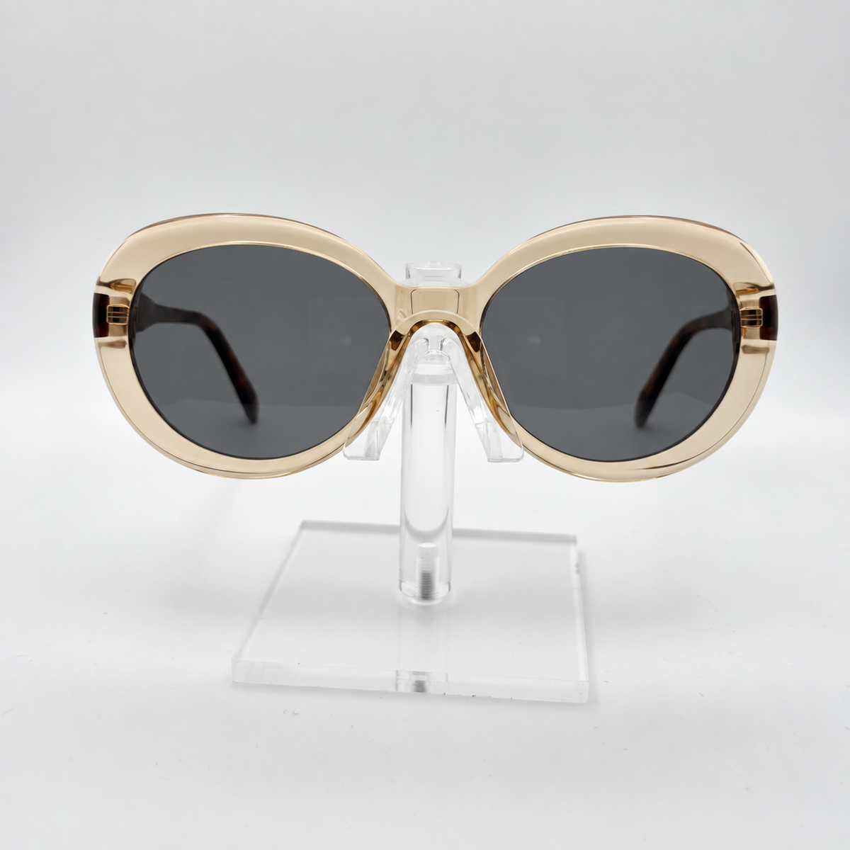 Oversized oval sunglasses with a gold-tone frame and dark gray lenses, displayed on a clear acrylic stand against a light gray background.