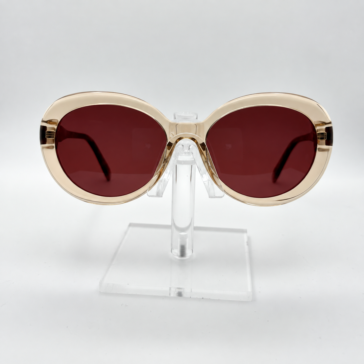 Oversized cat-eye sunglasses with a translucent champagne-gold frame and dark reddish-brown lenses, displayed on a clear acrylic stand against a light gray background.