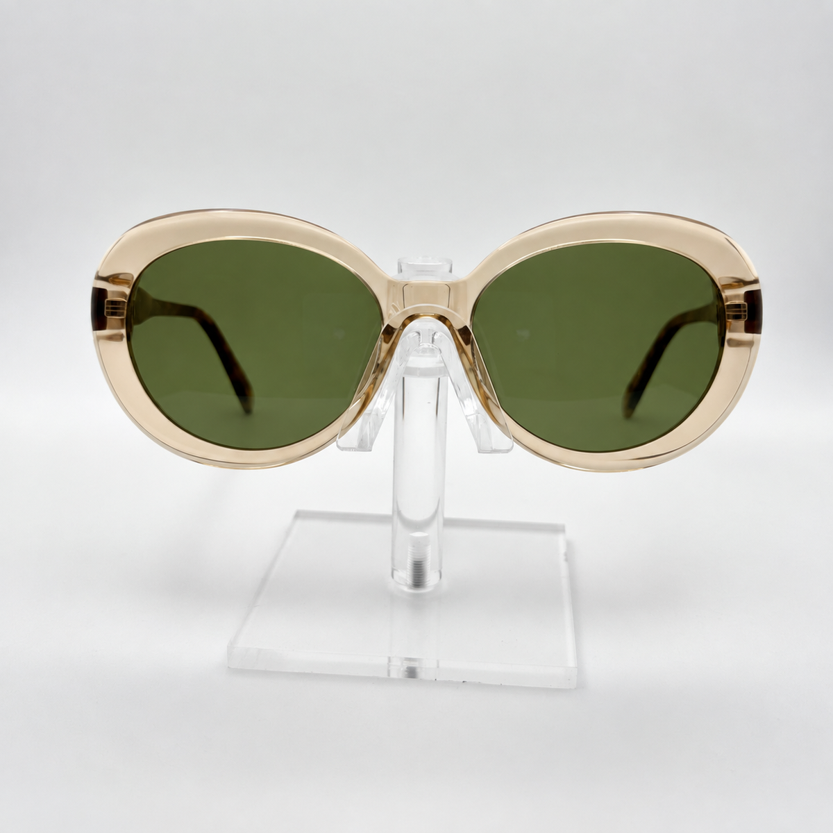Oversized oval sunglasses with a translucent champagne-gold frame and dark green lenses, displayed on a clear acrylic stand against a light gray background.