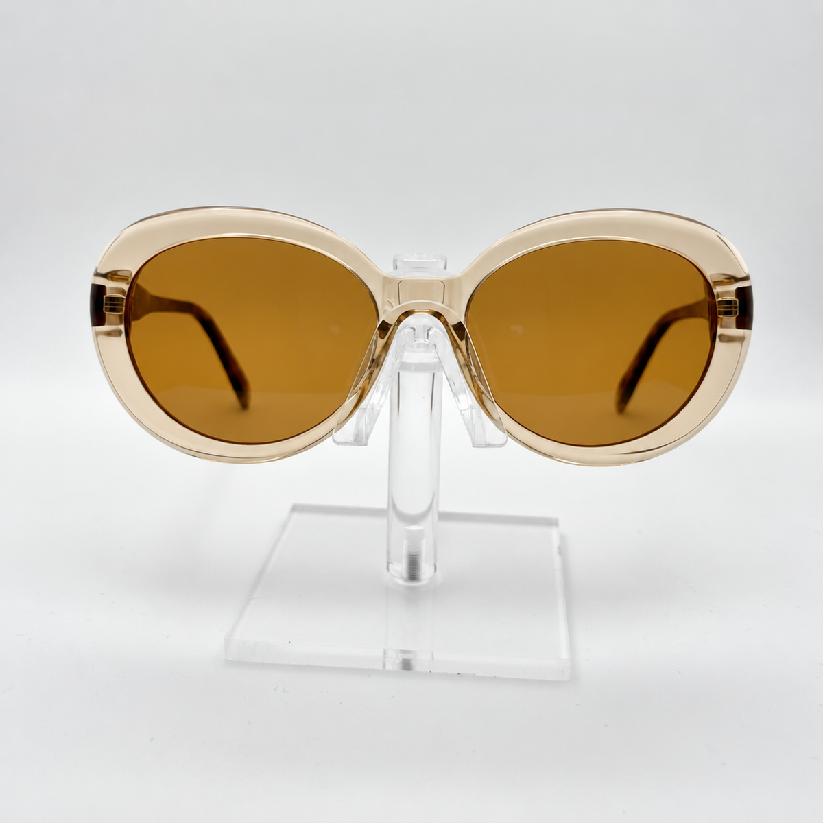 Oversized cat-eye sunglasses with translucent beige frames and warm brown lenses, displayed on a clear acrylic stand against a white background.