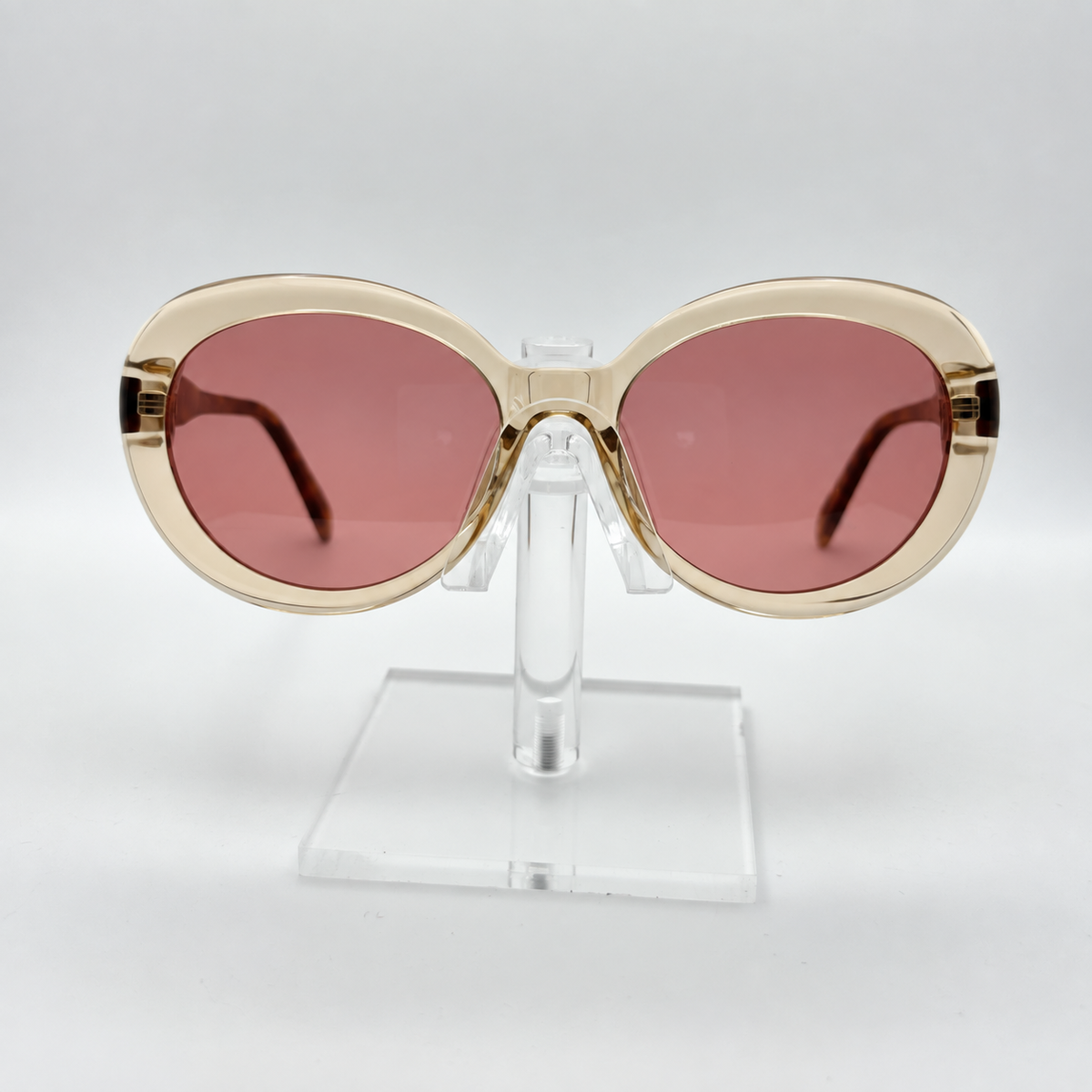 Oversized cat-eye sunglasses with a translucent beige frame and pink-tinted lenses, displayed on a clear acrylic stand against a light gray background.