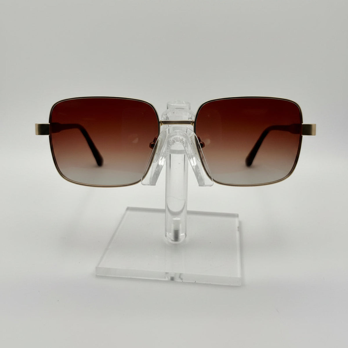 Square aviator-style sunglasses with brown gradient lenses and gold metal frames, displayed on a clear acrylic stand against a white background.