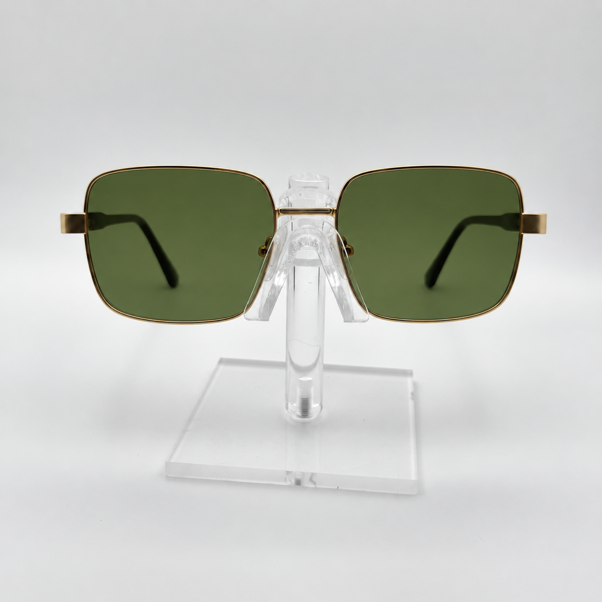 Square sunglasses with olive green lenses and gold metal frames, displayed on a clear acrylic stand against a light gray background.