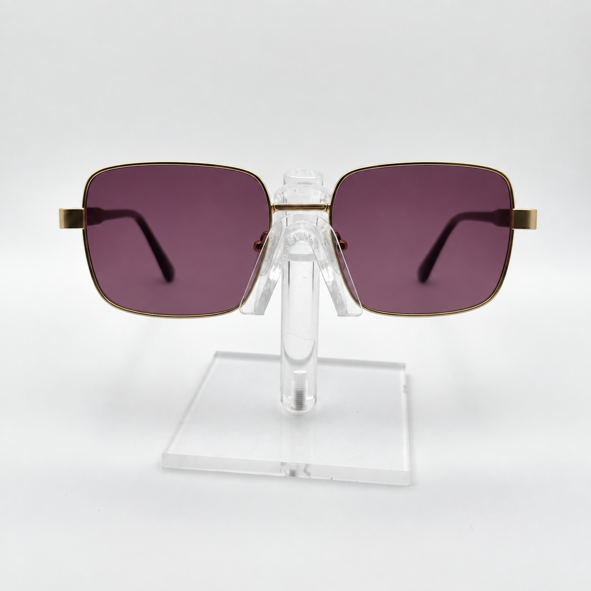 Square sunglasses with dark purple lenses and gold metal frames, displayed on a clear acrylic stand against a white background.
