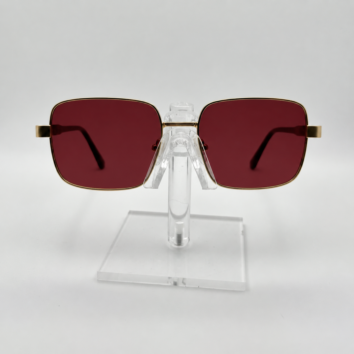 Square sunglasses with dark red lenses and gold metal frames, displayed on a clear acrylic stand against a light gray background.