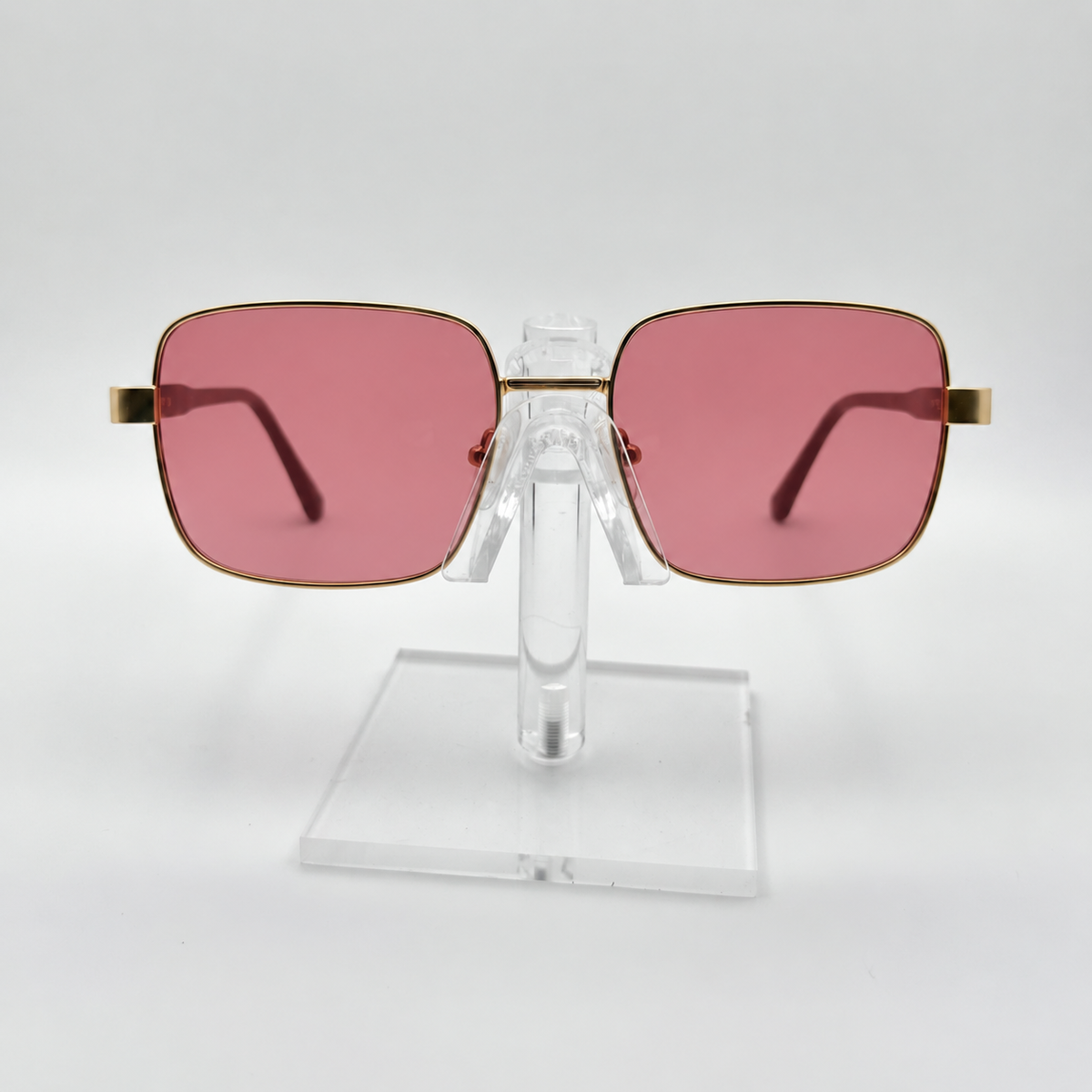 Square sunglasses with pink tinted lenses and gold metal frames, displayed on a clear acrylic stand against a light gray background.