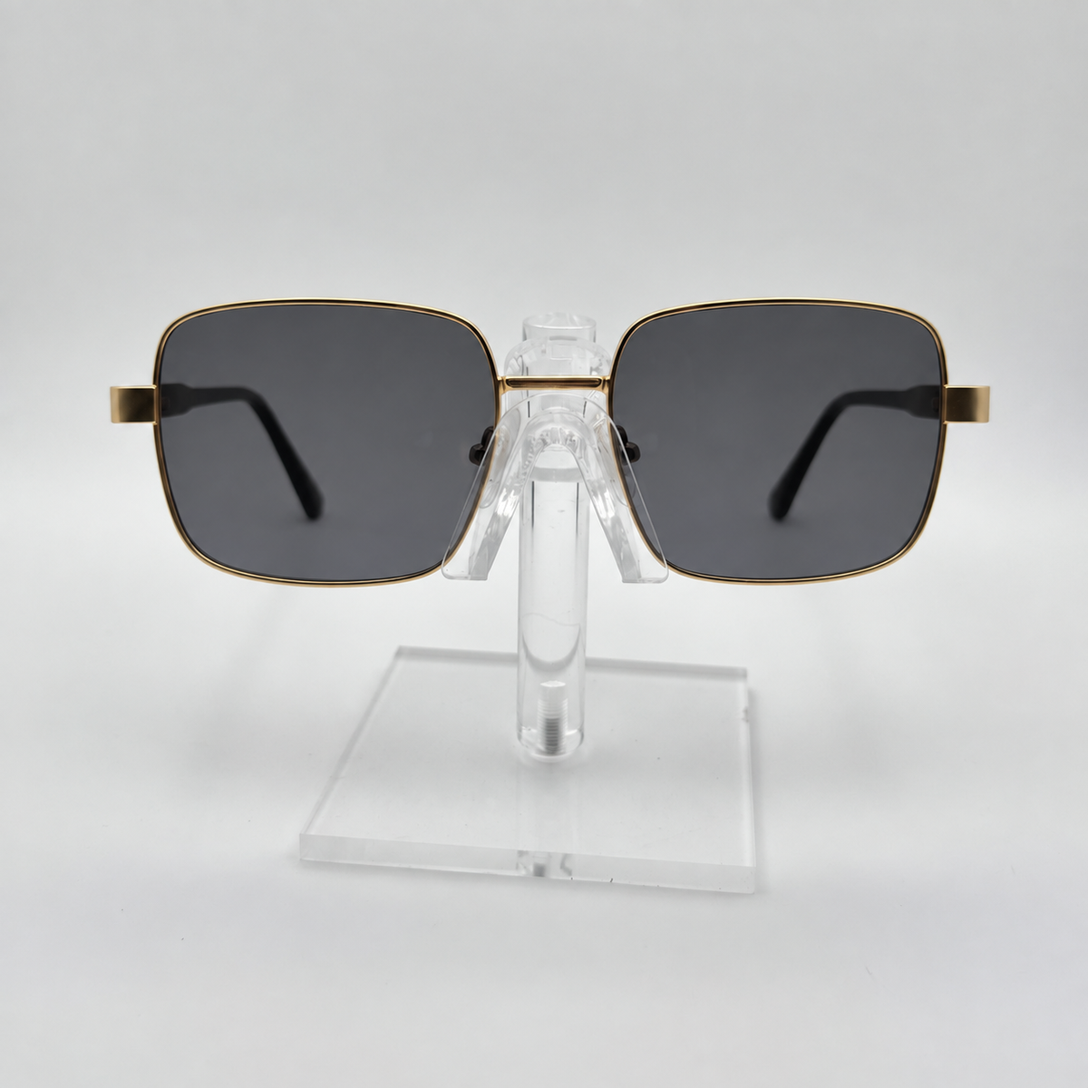 Square metal-framed sunglasses with dark gray lenses and gold-toned rims, displayed on a clear acrylic stand against a light gray background.