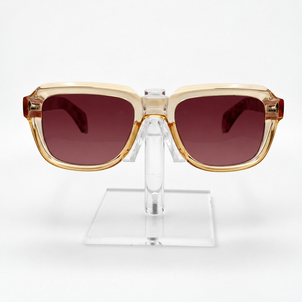 Square sunglasses with a translucent beige frame, gold-toned trim, and dark reddish-brown lenses, displayed on a clear acrylic stand against a white background.