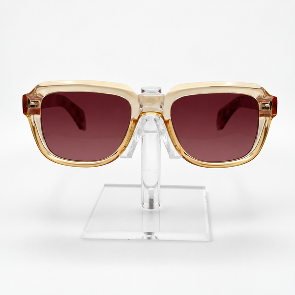 Square sunglasses with a translucent beige frame, gold-toned trim, and dark reddish-brown lenses, displayed on a clear acrylic stand against a white background.