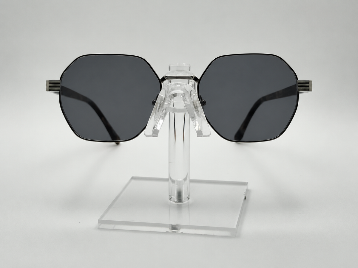 Oversized square sunglasses with dark gray lenses and black temples, displayed on a clear acrylic stand against a light gray background.