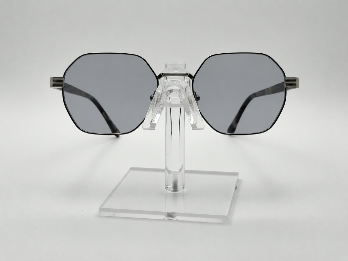 Oversized square sunglasses with dark gray lenses and black metal frames, displayed on a clear acrylic stand against a light gray background.