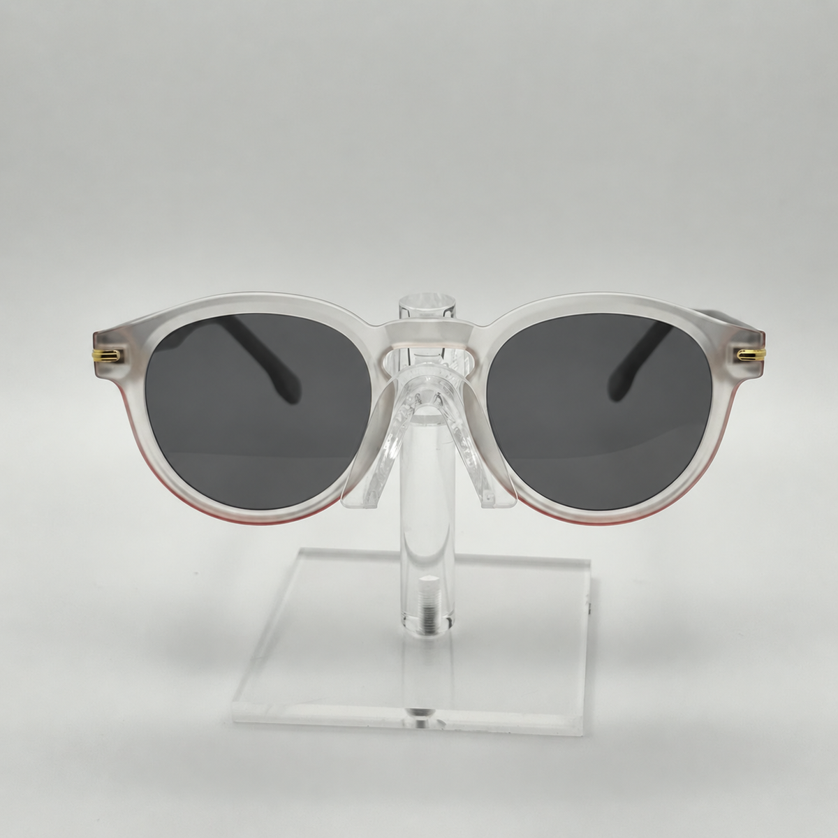 Round sunglasses with translucent white frames, dark gray lenses, and small gold metal accents on the temples, displayed on a clear acrylic stand against a light gray background.