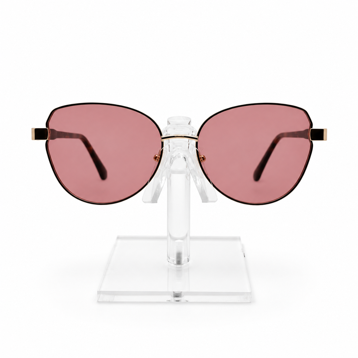 Oversized cat-eye sunglasses with dusty rose pink lenses and gold metal frames, displayed on a clear acrylic stand against a white background.