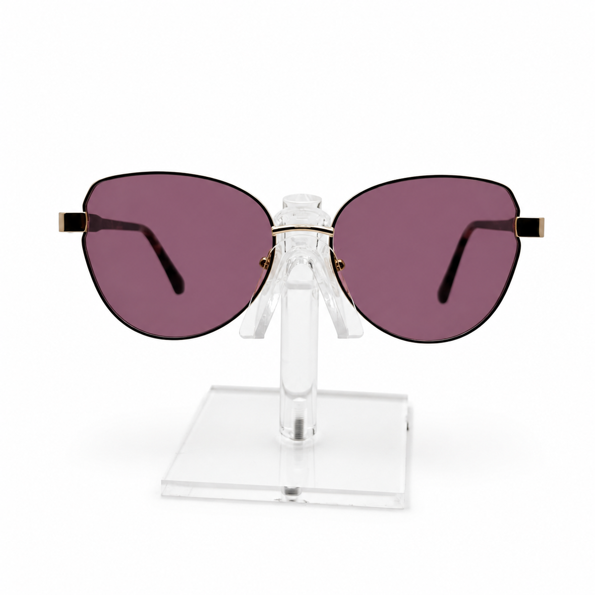 Oversized cat-eye sunglasses with dark purple lenses and gold metal hinges, displayed on a clear acrylic stand against a white background.