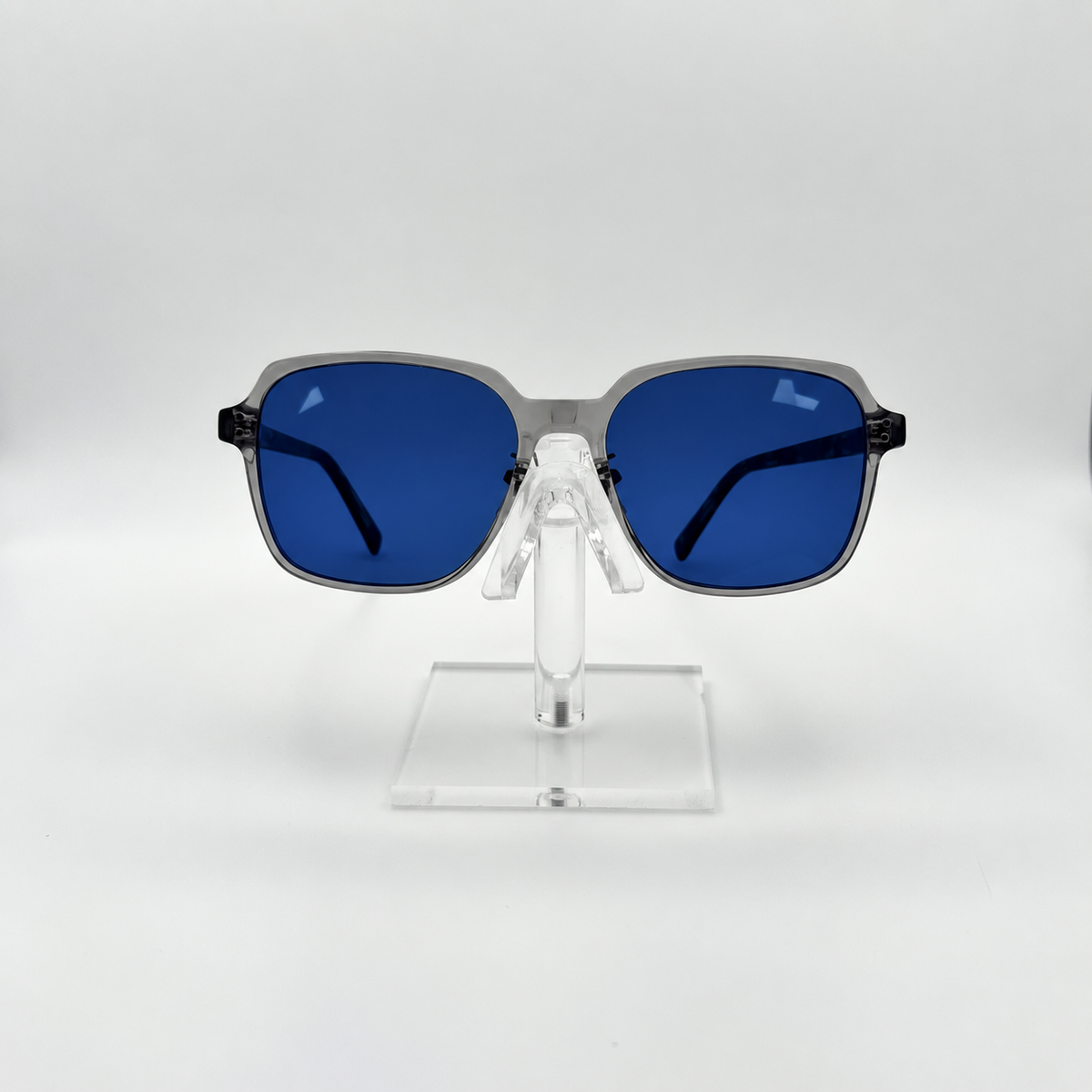 Square sunglasses with blue lenses and a gray frame, displayed on a clear acrylic stand against a white studio background.