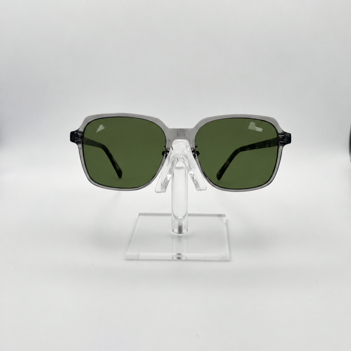 Square sunglasses with dark green lenses and a translucent gray frame, displayed on a clear acrylic stand against a light gray background.