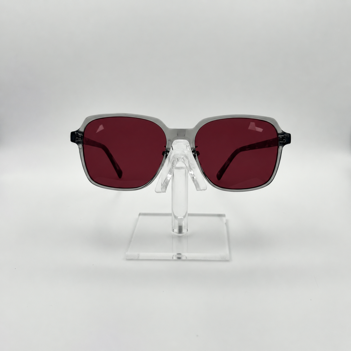 Square sunglasses with translucent gray frames and dark red lenses, displayed on a clear acrylic stand against a light gray background.