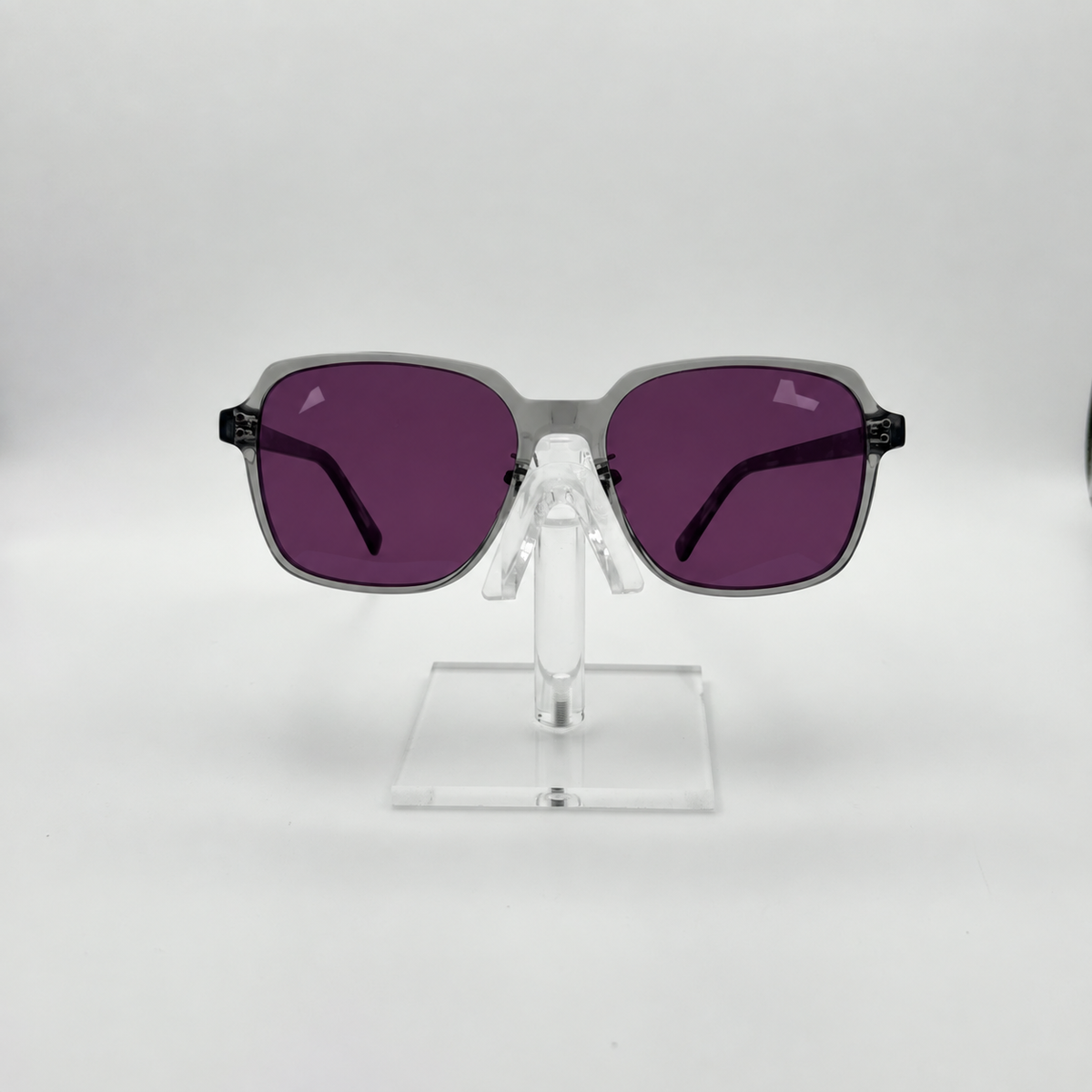 Square sunglasses with thick gray frames and dark purple lenses, displayed on a clear acrylic stand against a light gray background.