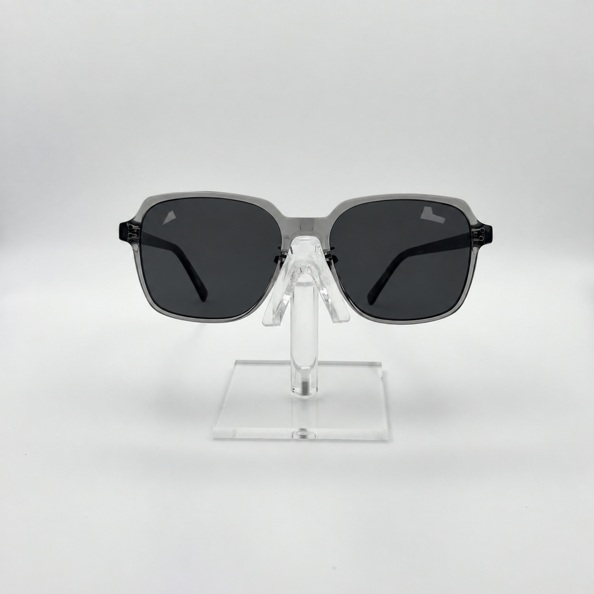 Square sunglasses with a translucent gray frame and dark gray lenses, displayed on a clear acrylic stand against a light gray background.