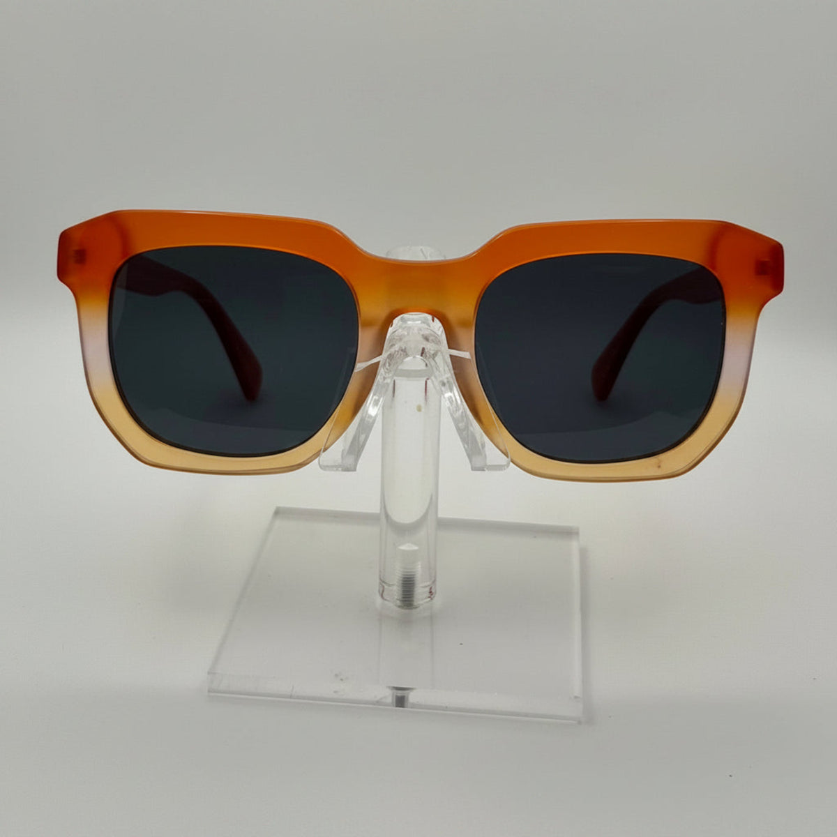 Square sunglasses with a translucent orange-to-tan gradient frame and dark lenses, displayed on a clear acrylic stand against a light gray background.