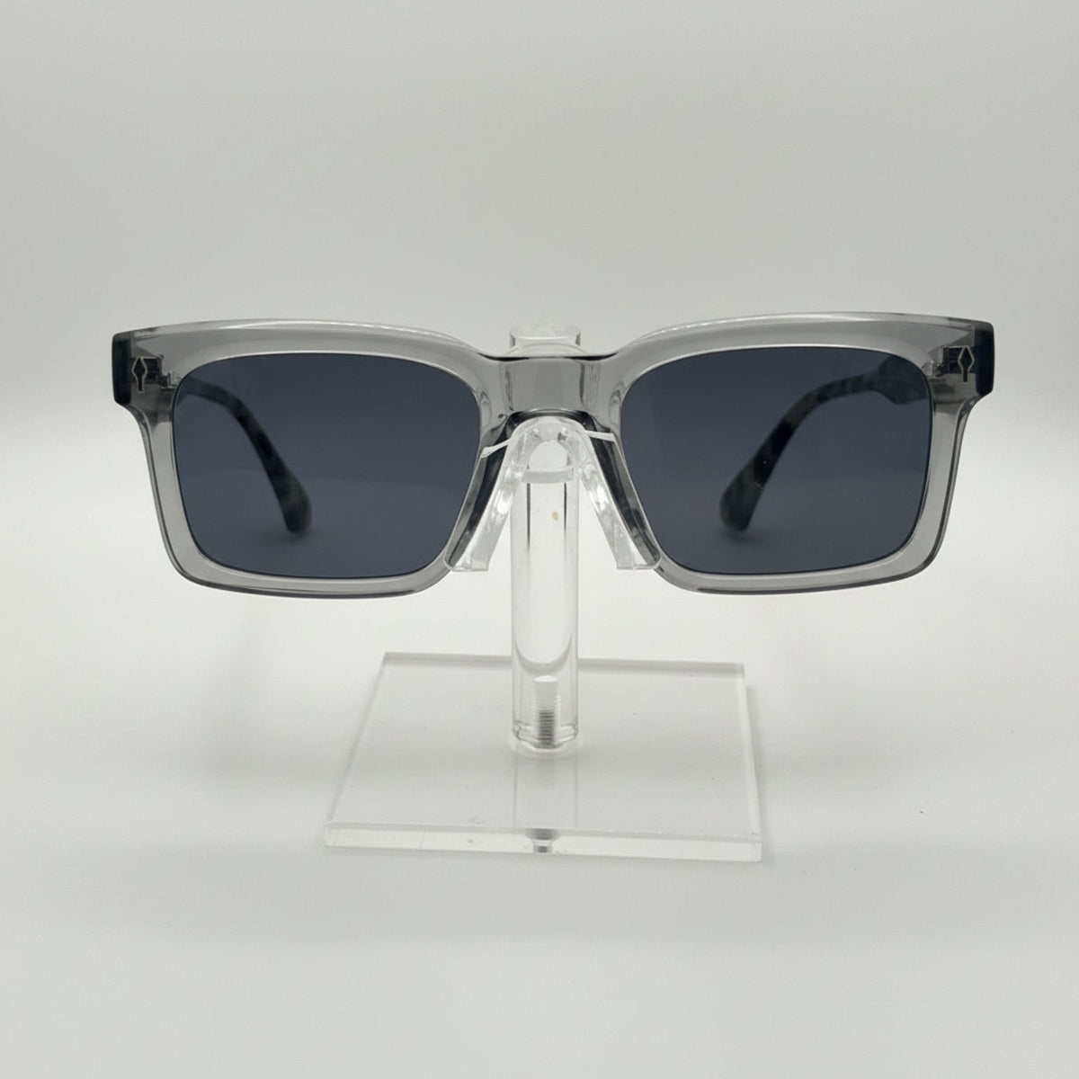 Square sunglasses with a translucent smoky gray frame and dark gray lenses, displayed on a clear acrylic stand against a light gray background.
