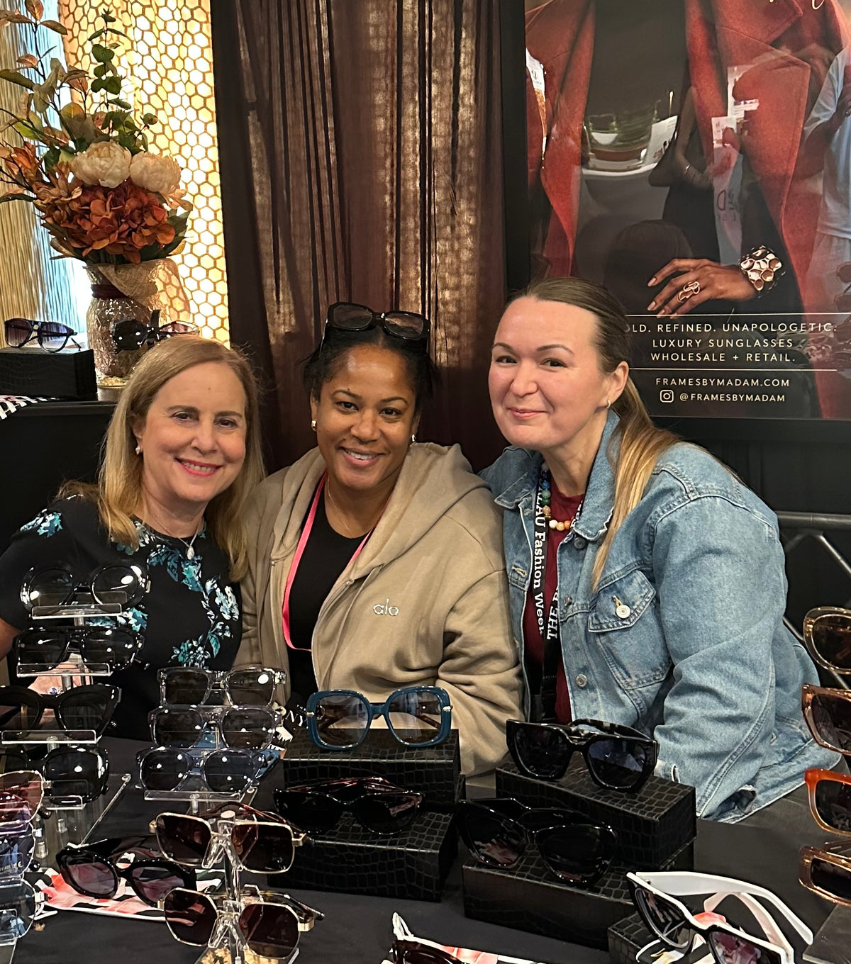 Three women smiling and posing together at a retail display table filled with sunglasses in black and leather cases. The setting has warm brown curtains and a decorative floral arrangement in the background.