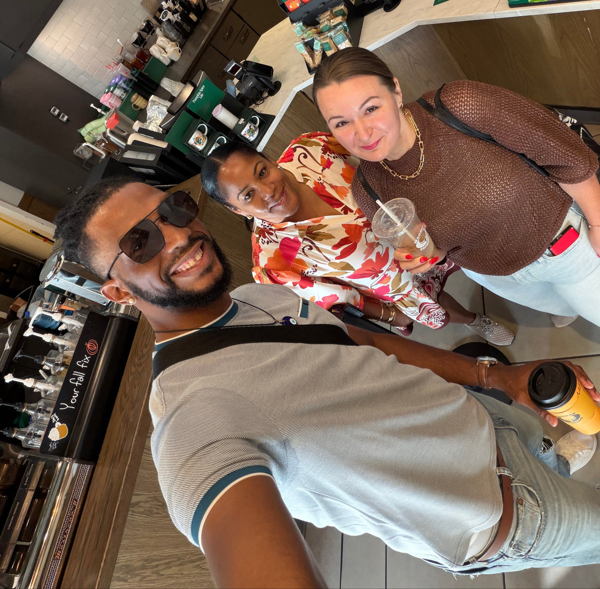 Three people taking a selfie inside a Starbucks coffee shop, with the counter, coffee machines, and shelves visible in the background.