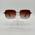 Square aviator-style sunglasses with brown gradient lenses and gold metal frames, displayed on a clear acrylic stand against a white background.