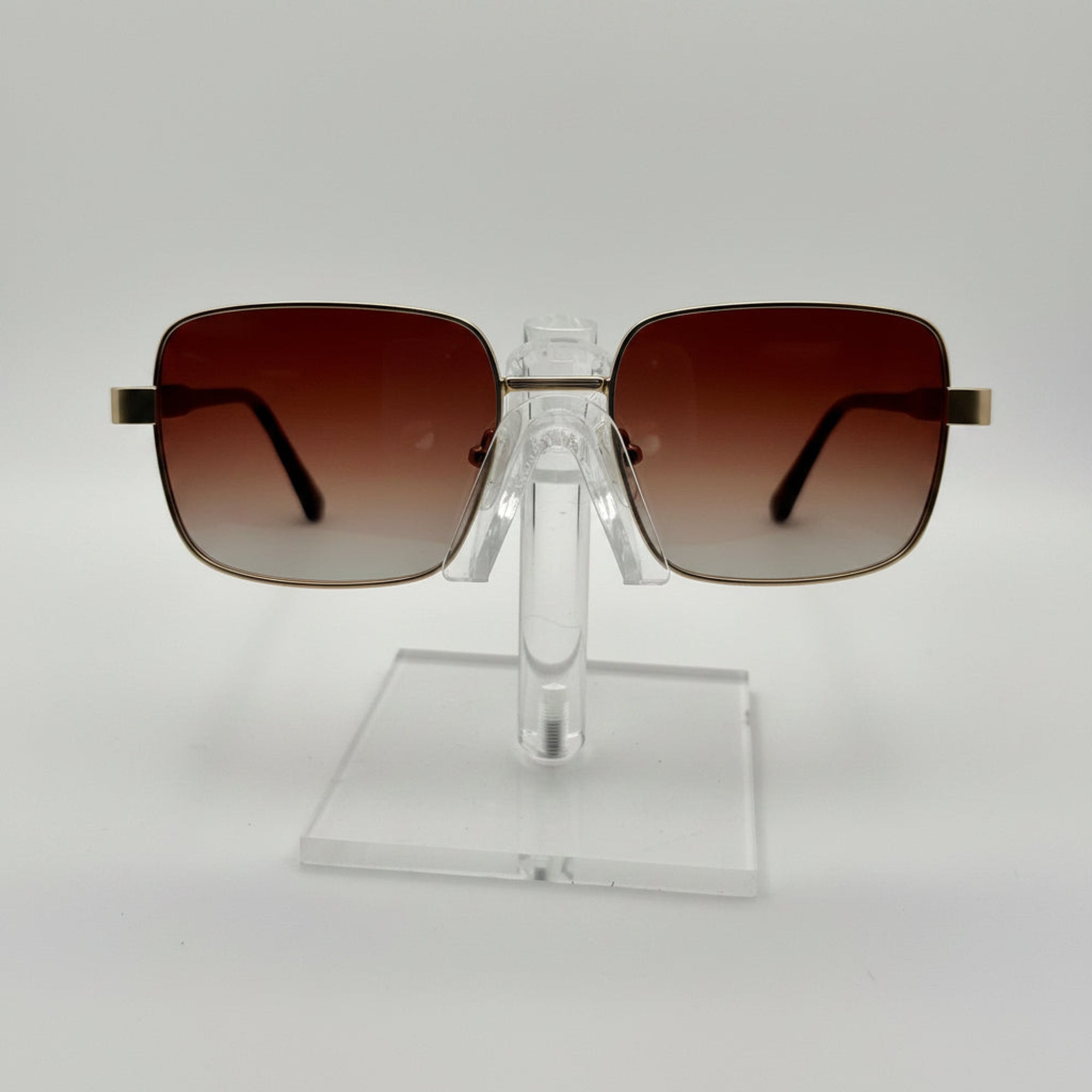 Square aviator-style sunglasses with brown gradient lenses and gold metal frames, displayed on a clear acrylic stand against a white background.