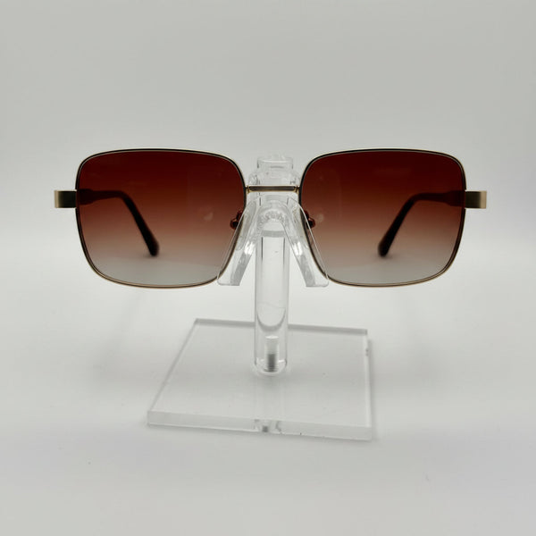 Square aviator-style sunglasses with brown gradient lenses and gold metal frames, displayed on a clear acrylic stand against a white background.