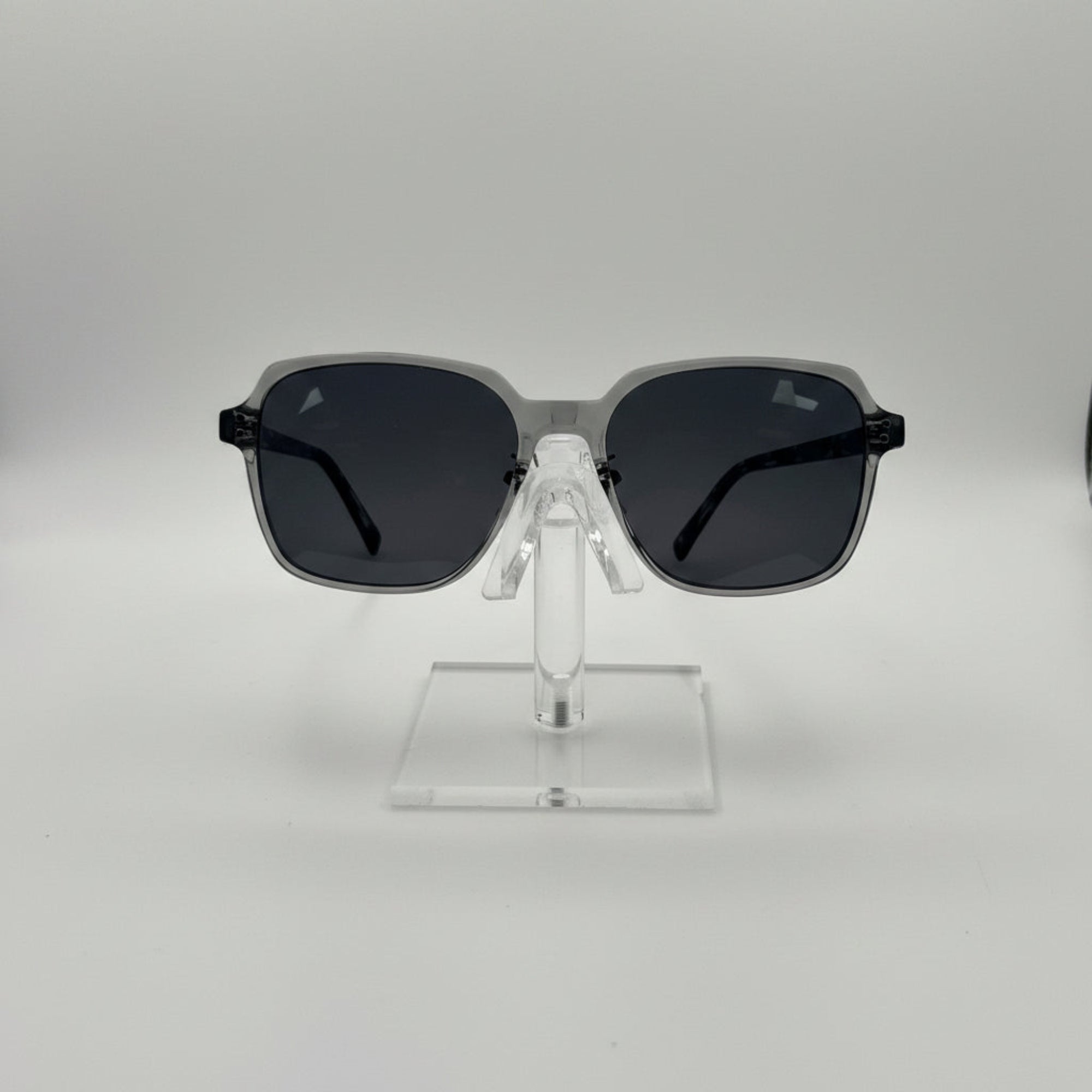 Oversized square sunglasses with a translucent smoky gray frame and dark gray lenses, displayed on a clear acrylic stand against a light gray background.