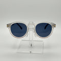 Round sunglasses with a translucent white frame and dark blue lenses, displayed on a clear acrylic stand against a light gray background.