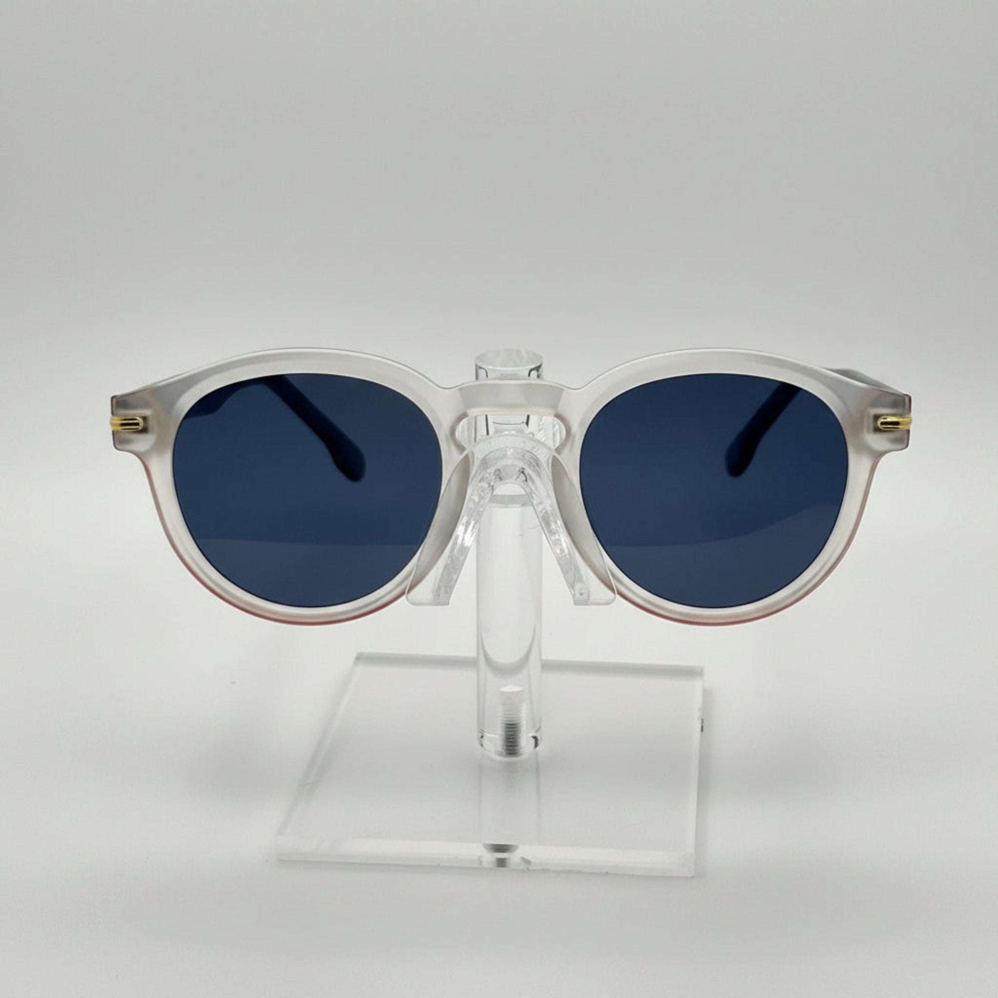 Round sunglasses with a translucent white frame and dark blue lenses, displayed on a clear acrylic stand against a light gray background.