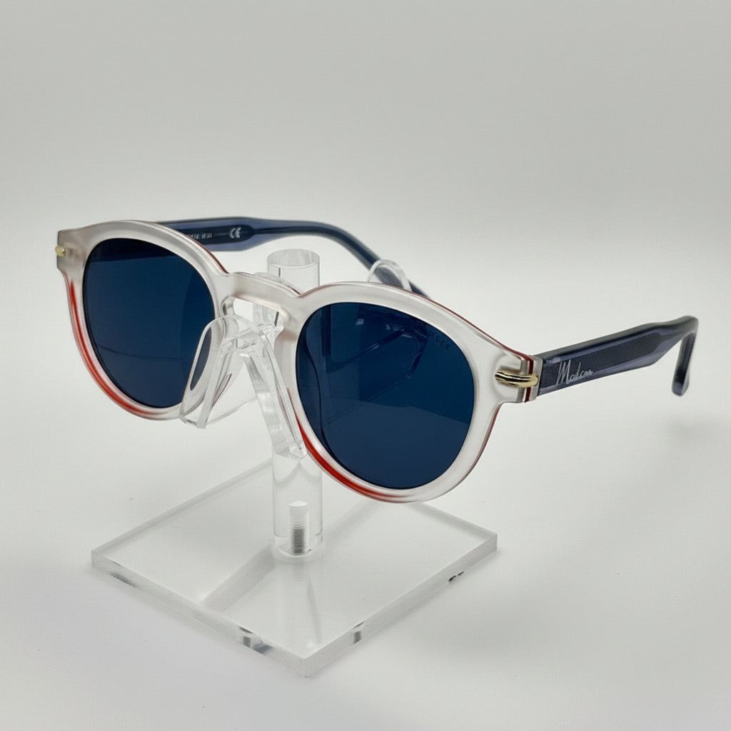 A pair of round sunglasses with a translucent white frame, dark blue lenses, and a thin red accent line along the inner edge of the frame, displayed on a clear acrylic stand against a white background.