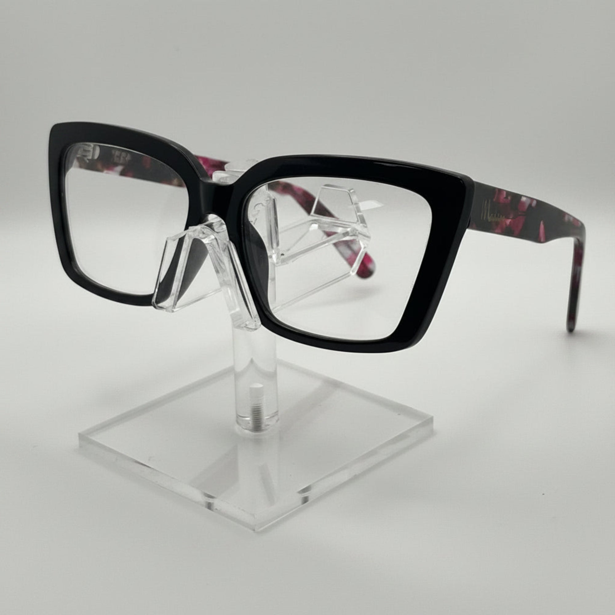 Black square eyeglasses with a tortoiseshell pattern on the temples, displayed on a clear acrylic stand against a light gray background.