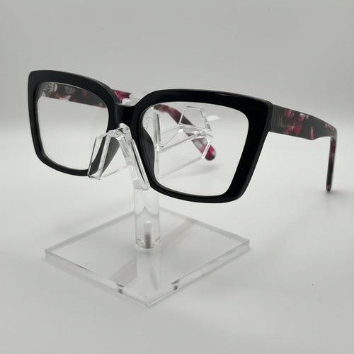 Black square eyeglasses with a tortoiseshell pattern on the temples, displayed on a clear acrylic stand against a light gray background.