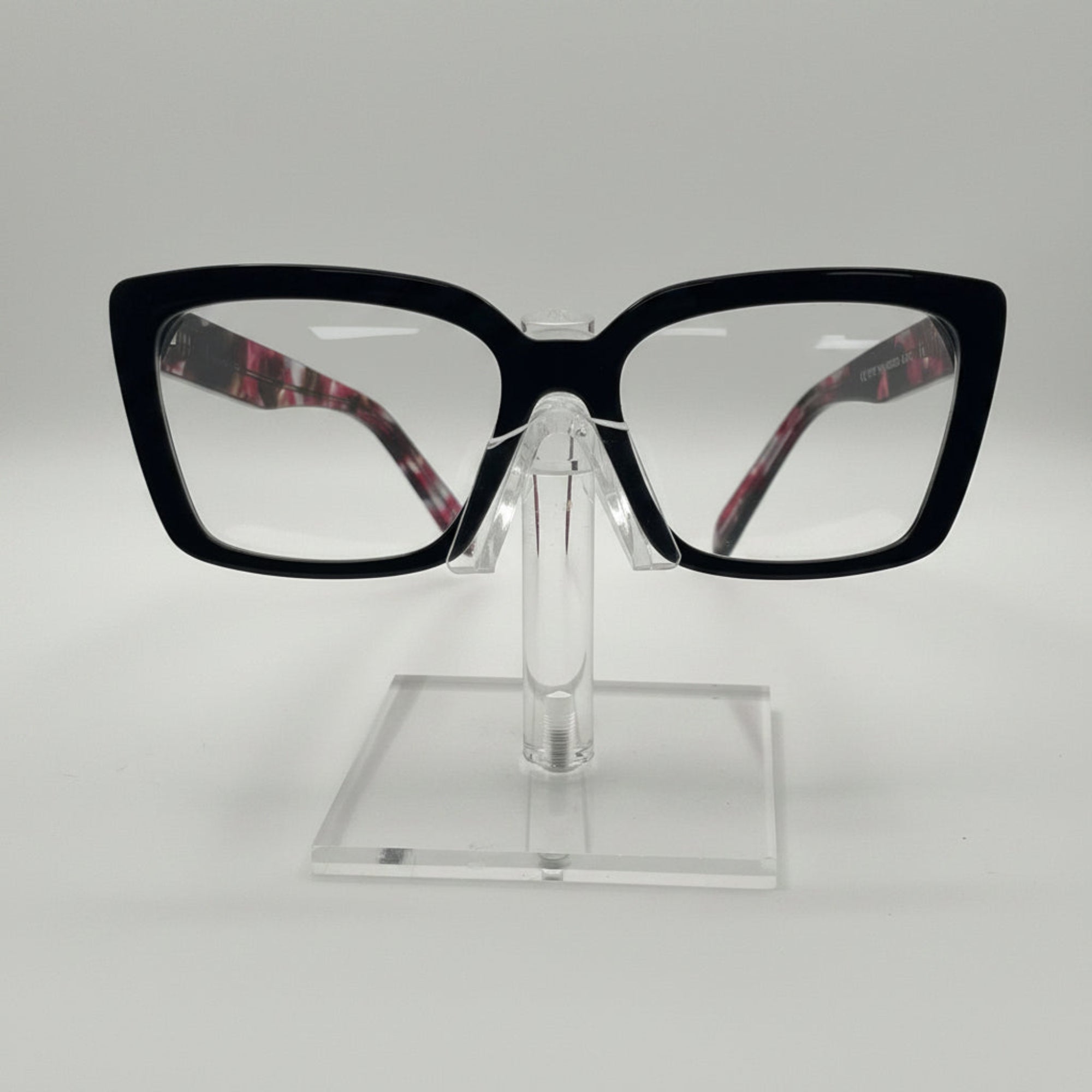 Black cat-eye eyeglasses with clear lenses and patterned inner temples, displayed on a clear acrylic stand against a white background.