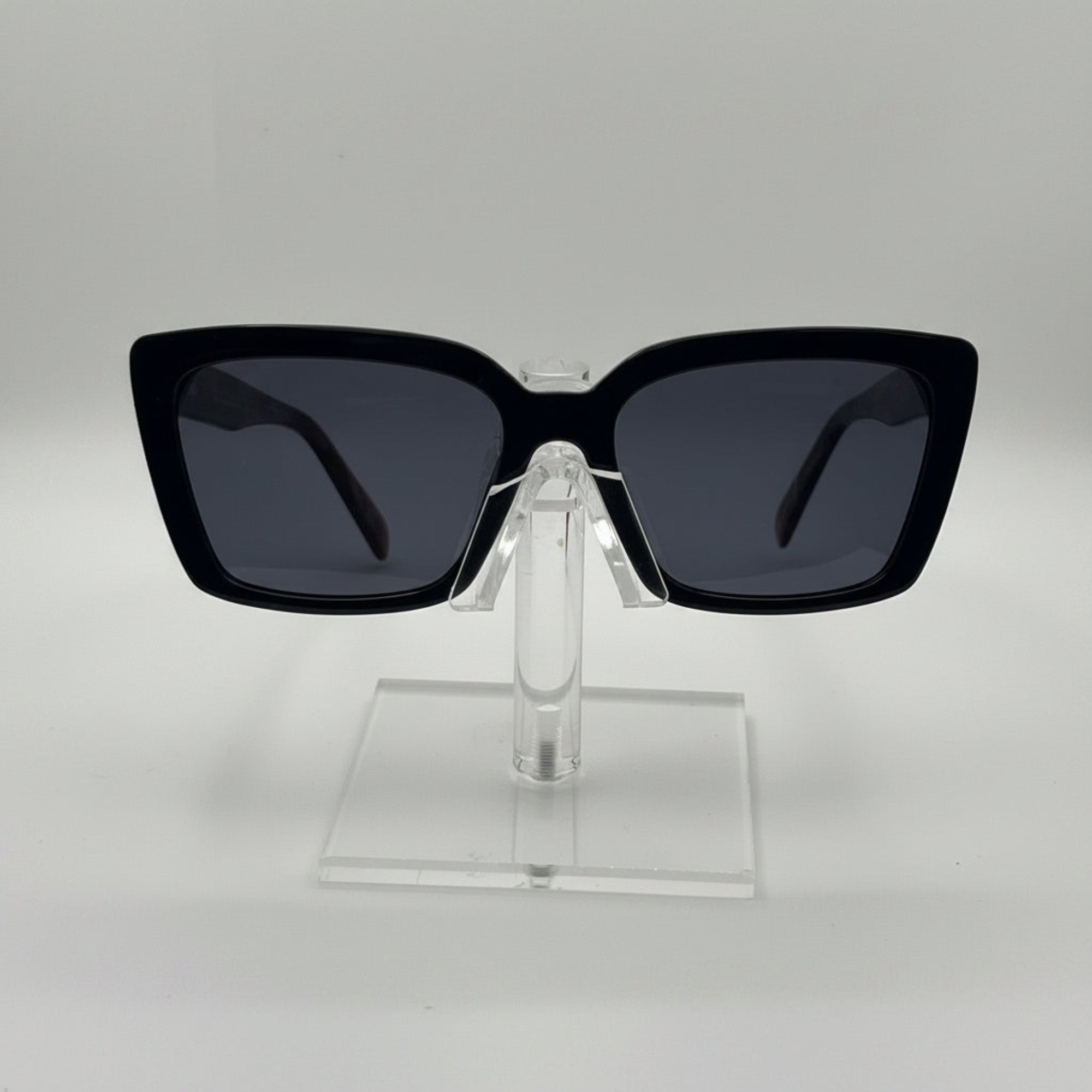 Black square sunglasses with dark lenses displayed on a clear acrylic stand against a light gray background.