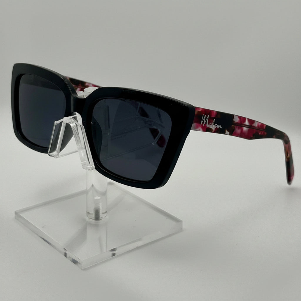 A pair of square sunglasses with a thick black front frame and dark lenses, featuring patterned arms in red and black, displayed on a clear acrylic stand against a light gray background.
