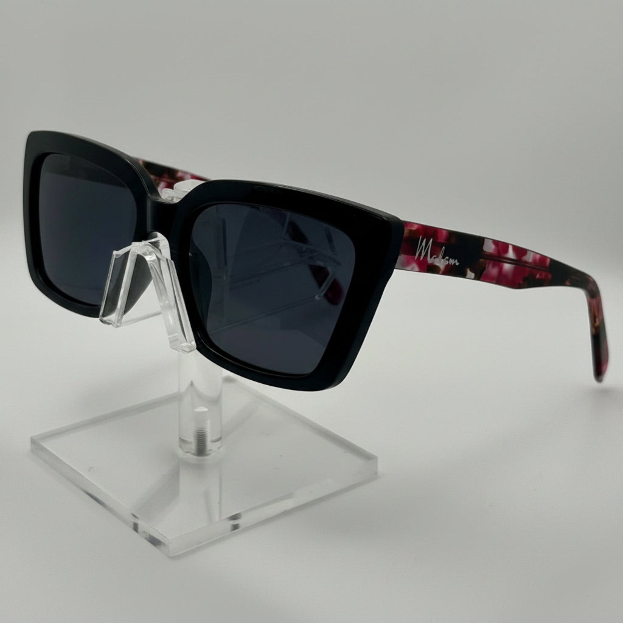 A pair of square sunglasses with a thick black front frame and dark lenses, featuring patterned arms in red and black, displayed on a clear acrylic stand against a light gray background.