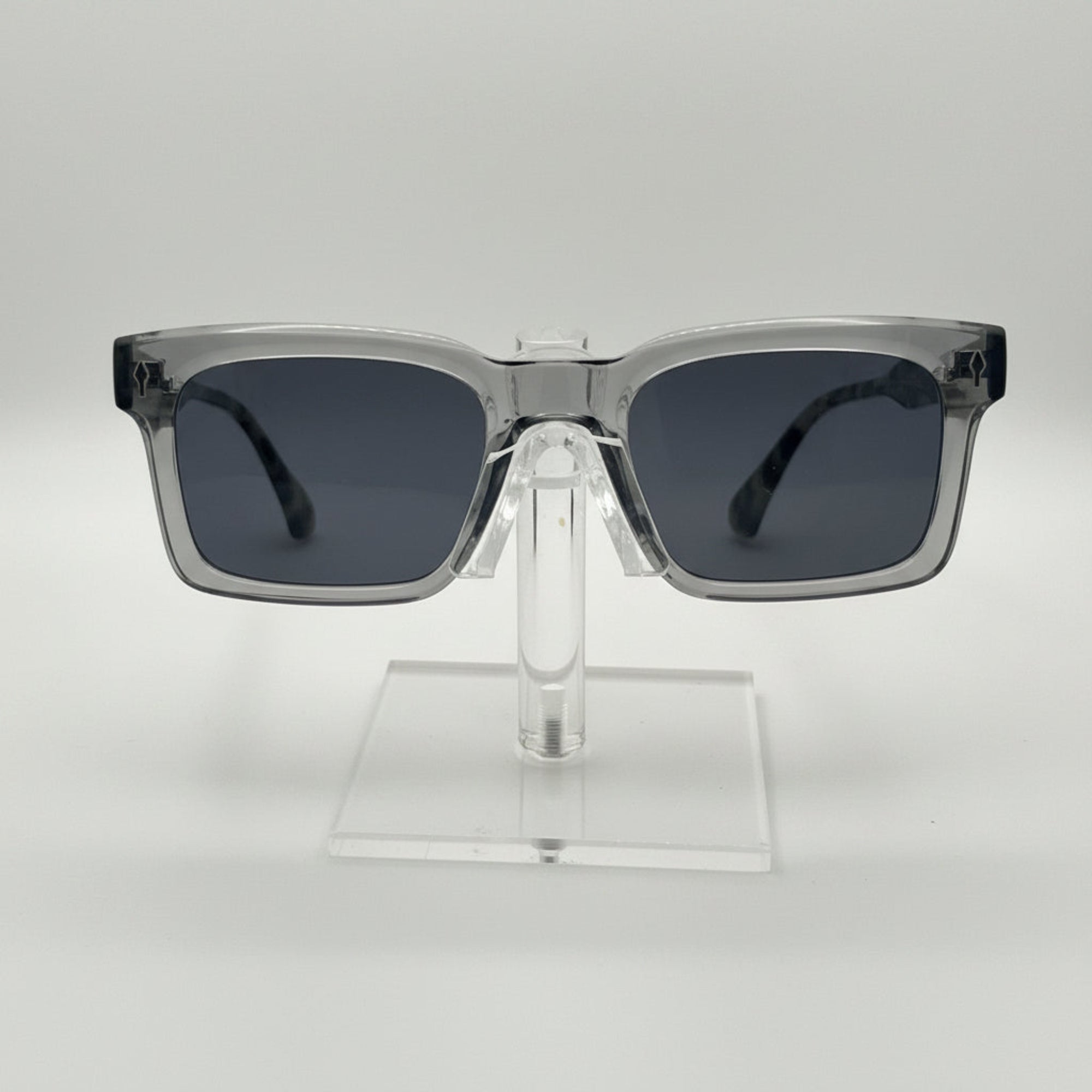 Square sunglasses with a translucent smoky gray frame and dark gray lenses, displayed on a clear acrylic stand against a light gray background.