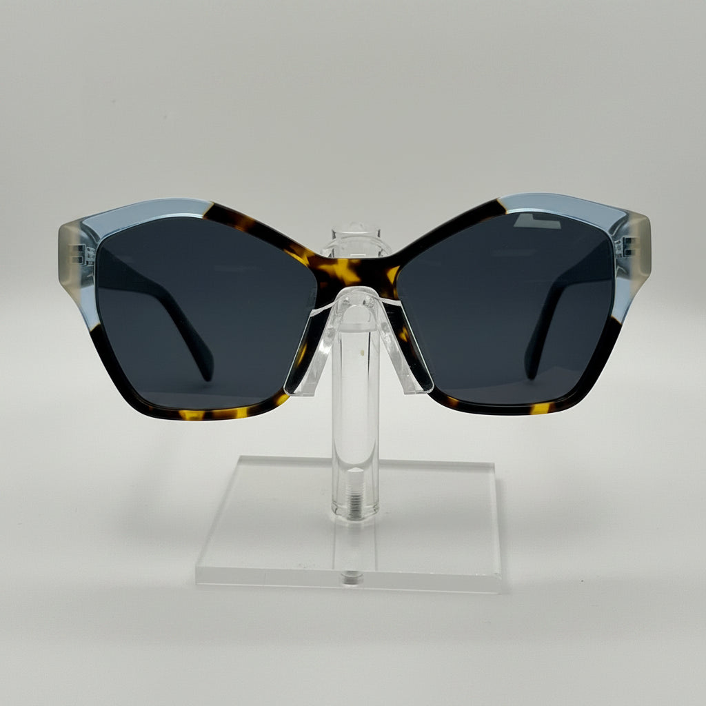 Oversized square sunglasses with dark lenses, a black and brown tortoiseshell frame, and translucent light blue temples, displayed on a clear stand against a light gray background.