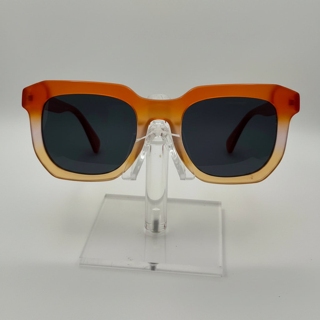 Square sunglasses with a translucent orange-to-tan gradient frame and dark lenses, displayed on a clear acrylic stand against a light gray background.